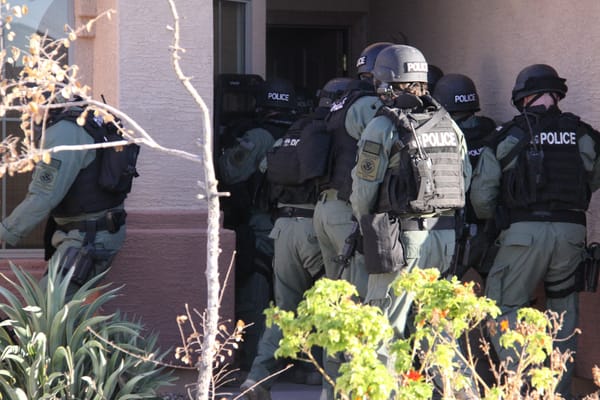 ICE officers in tactical combat gear with vests that read "Police" on the back gather outside the door of a building preparing to enter.