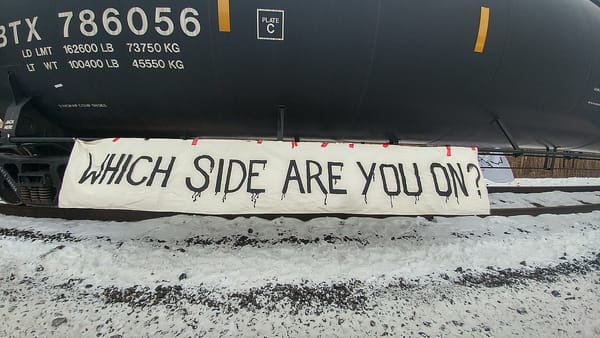 A sign reading "WHICH SIDE ARE YOU ON" is displayed below a tanker car on an train carrying liquified natural gas.