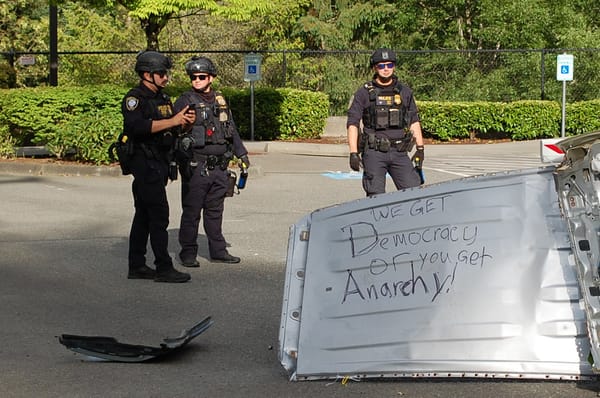 Homeland Security agents in black tactical gear stand in front of a metal barricade placed by protesters that reads: We get democracy or you get anarchy!