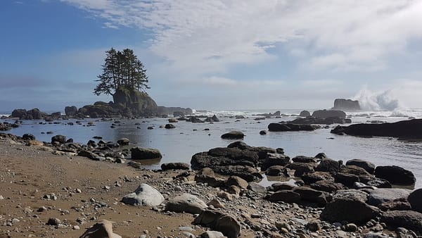 A wave crashes on a small island offshore of a rocky beach. In the distance another small island is topped by several Sitka Spruce.