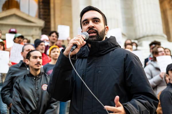 New York mayor candidate Zohran Mamdani in a black rain jacket, speaks into a microphone to a crowd of protesters opposing fascism.