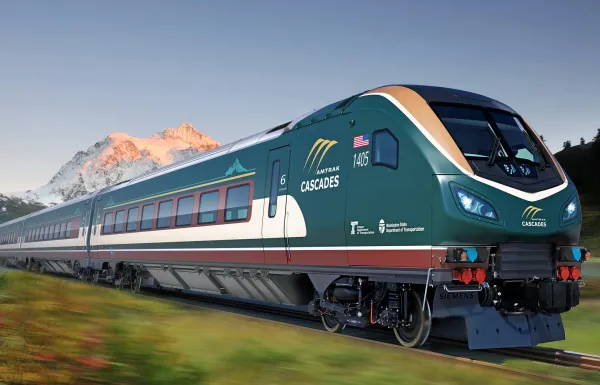 A sleek forest green passenger train with the Amtrak Cascades logo speeds down a track with Mount Rainier in the background.