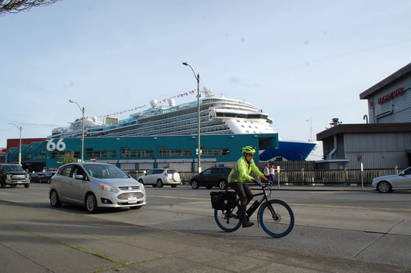 On the Seattle waterfront, a large cruise ship is docked at pier 66 as a car and a cyclist pass in front of it.