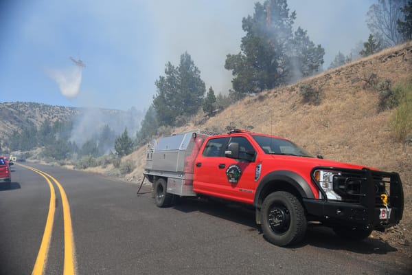 A forest fighting pickup truck is seen on the side of a highway as a helicopter drops water on a fire among scattered pine trees.