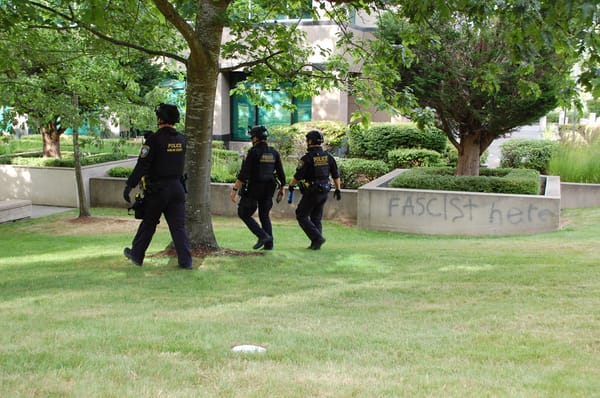 Three DHS officers in black tactical gear and helmets walk across a lawn next to a wall spray-painted with the words "FASCIST HERE."