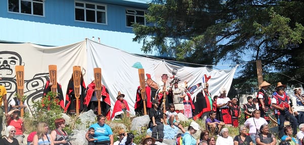Members of the Heiltsuk First Nation gather wearing traditional red and black shell blankets, cedar hats, and holding traditional canoe paddles.