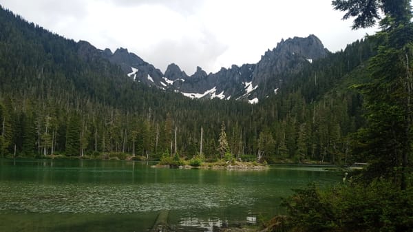 A mountain lake with green-tinted waters sits surrounded by forests of mountain hemlock and above it all, a sawtooth ridge of snowy mountains.