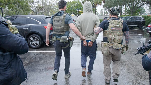 Officers of ICE detain a man, a person of color, in handcuffs, with another officer holding a military-stye assault rifle.
