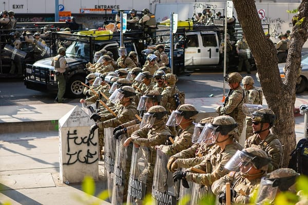 National guard troops with shields and helmet stand in a row in front of a trash can spray painted with the words Fuck ICE.