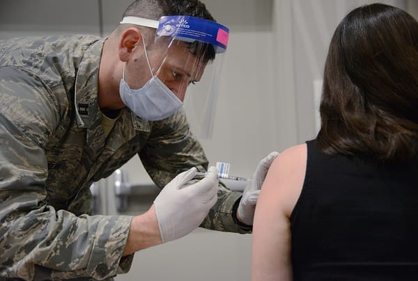 A man in camouflage fatigues and a face shield gives someone an injection.