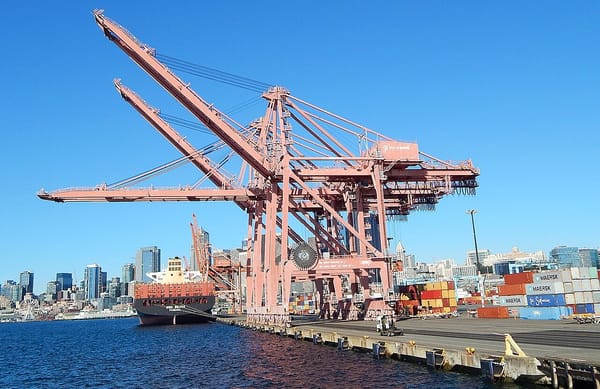 Large cranes load and unload containers above a cargo ship, with the Seattle skyine in the background.