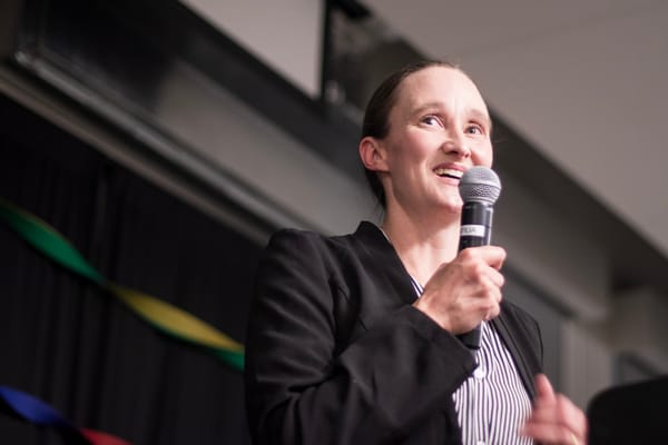 Katie Wilson wears a striped shirt and jacket and speakes into a microphone.