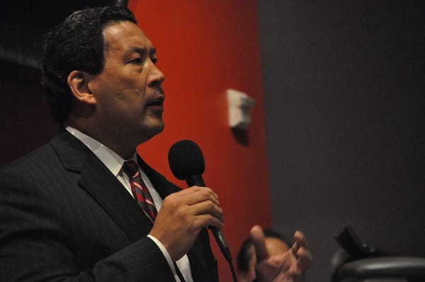 Bruce Harrell speaks into a microphone with a dark red wall behind him.