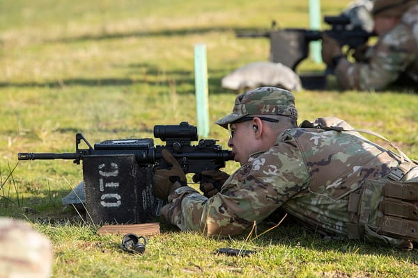 A soldier in camoflauge lying on the ground aims an assault rifle.