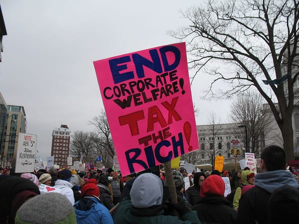 A crowd of people protesting, with one holding a sign that reads: End Corporate Welfare! Tax the Rich!
