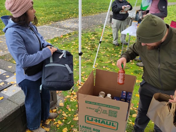 A volunteer puts a bottle of hot sauce into a box of dry goods as volunteers drop off food under rain canopies.