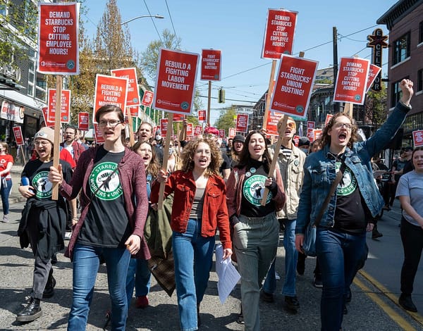 Starbucks workers in union t-shirts march down a street carrying signs that read Demand Starbucks Stop Cutting Employee Hours