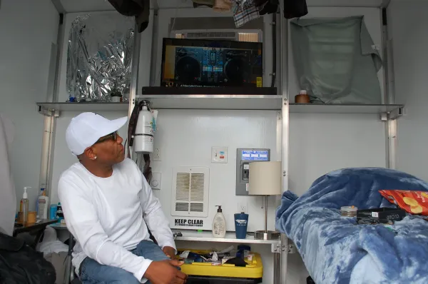 A man wearing a cap and sunglasses sits in a small home with a bed, lamp, TV and various personal items