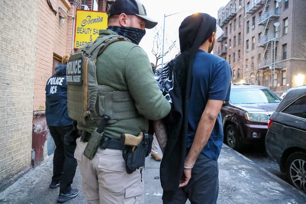 A federal agent with a gun and face mask puts handcuffs on a man on a city street.