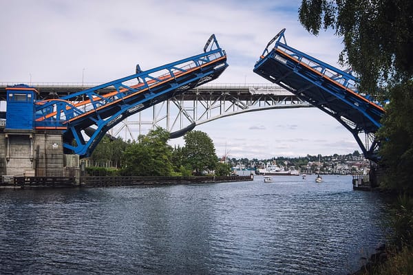 A blue drawbridge rises over the waters of Lake Union, with boats in the background