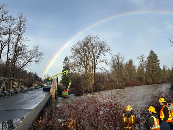 A rainbow shines over a highway bridge, with workers using a crane to pull debris from a flooded river