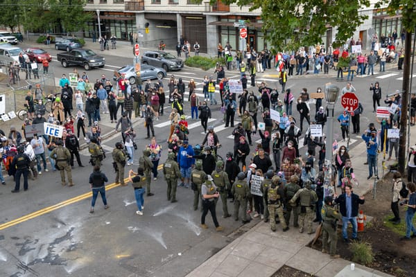 A crowd of protesters, seen from above, face down armed goon sin military gear. 