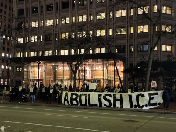 Protesters stand in front of an office building at night holding signs and a large banner that reads: Abolish ICE