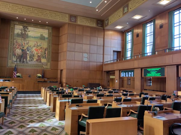Desks and chairs in the art deco Oregon house chamber, with a mural of pioneers behind the dais.
