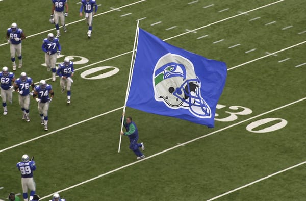 A man runs across a football field with a Seahawks flag as players look on.