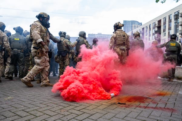 Agents in tactical gear and gas masks look menacing as a cannister emits pink-red smoke.