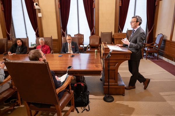 A man in a suit stand behind a podium whil members of the press sit at a table with cellphones and notebooks.