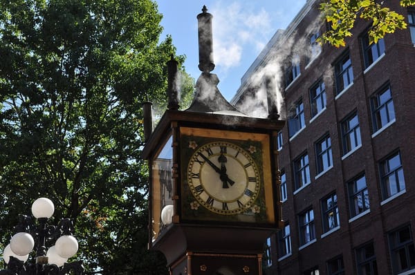 An old fashioned outdoor clock emits steam from an exhaust whistle.