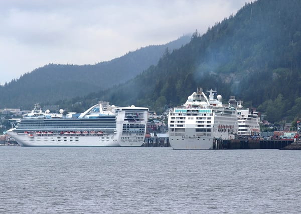 Three massive cruise ships docked at a port surrounded by forested mountains spew pollution from their smoke stacks